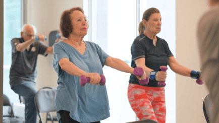 Residents focus intently as they lift hand weights during a group fitness class at Mirabelle, an active senior living community in Miami, FL with wellness programs designed to support strength and brain health.