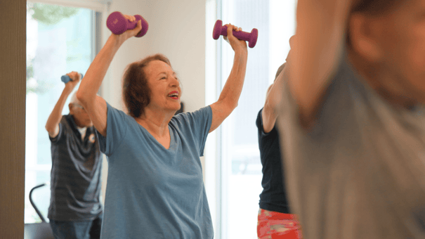 A resident laughs joyfully while raising hand weights overhead during a group fitness class at Mirabelle, an active senior living community in Miami, FL where wellness programs help residents thrive every day.