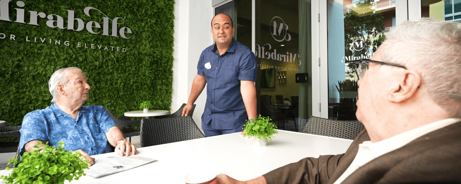 A staff member in a blue uniform speaks with two elderly male residents seated at an outdoor patio table at Mirabelle senior living community, with a green living wall and the Mirabelle logo visible in the background.