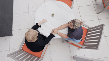 Aerial view of two women sitting across from each other at a round outdoor table, holding hands in a gesture of comfort or support. One woman, wearing a black top with short blonde hair, and another wearing a dark top and blue scrubs with a gold watch, are seated in wooden chairs with orange cushions on a light stone patio.