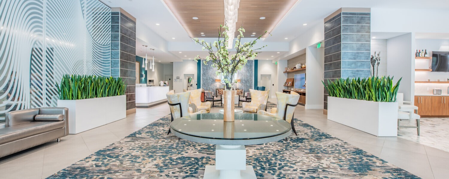 Amplio y luminoso salón de entrada en Mirabelle, con centro floral, tapete decorativo y vegetación exuberante, ideal para el bienestar diario de nuestra comunidad de senior living en Miami, FL.