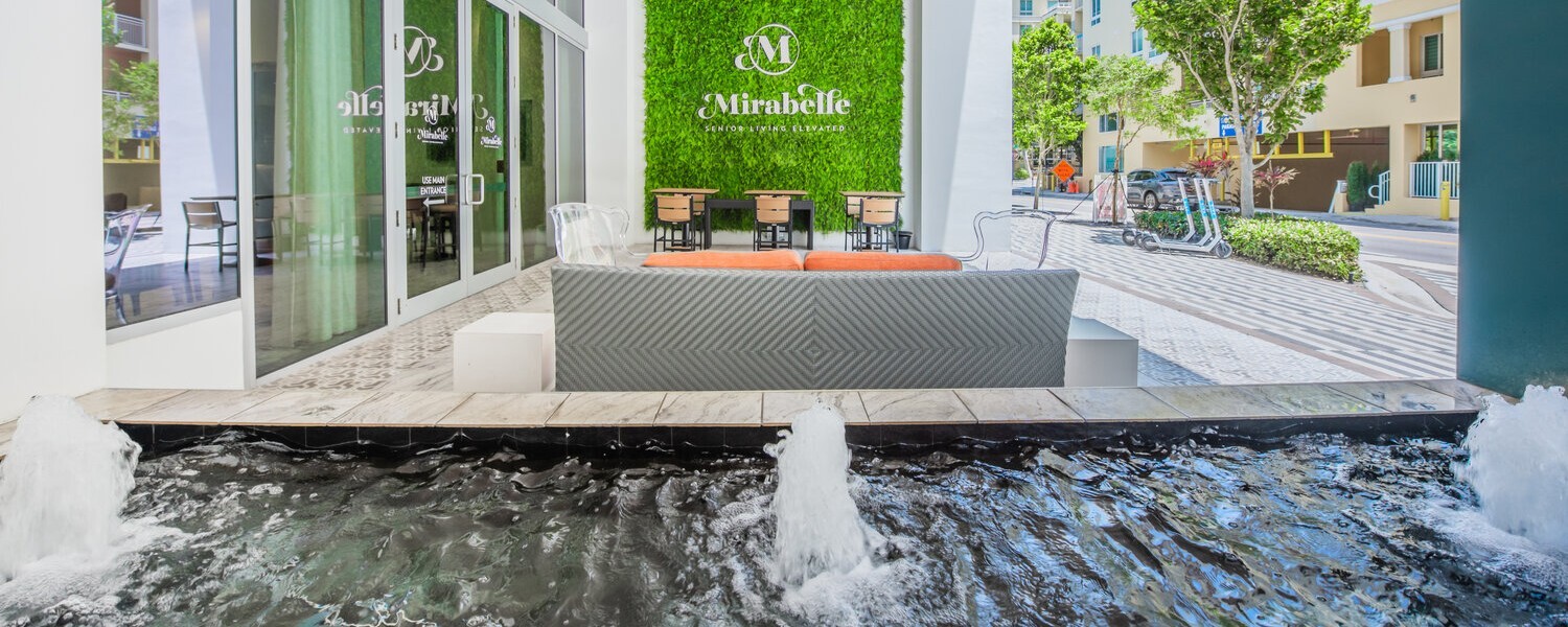 The sunlit outdoor entrance of Mirabelle senior living in Miami, FL, featuring a living green wall display, the community's logo, and water feature welcoming residents and visitors.
