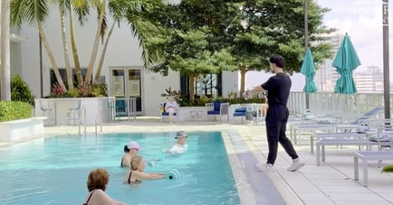 A rooftop pool deck where a fitness instructor in a black outfit stands poolside leading a water aerobics class. Four women stand in the pool holding foam dumbbells, wearing swim caps or sun hats. Behind them, lounge chairs, teal umbrellas, palm trees, and city high-rises are visible under a bright sky, with two seated onlookers in the background near the building.