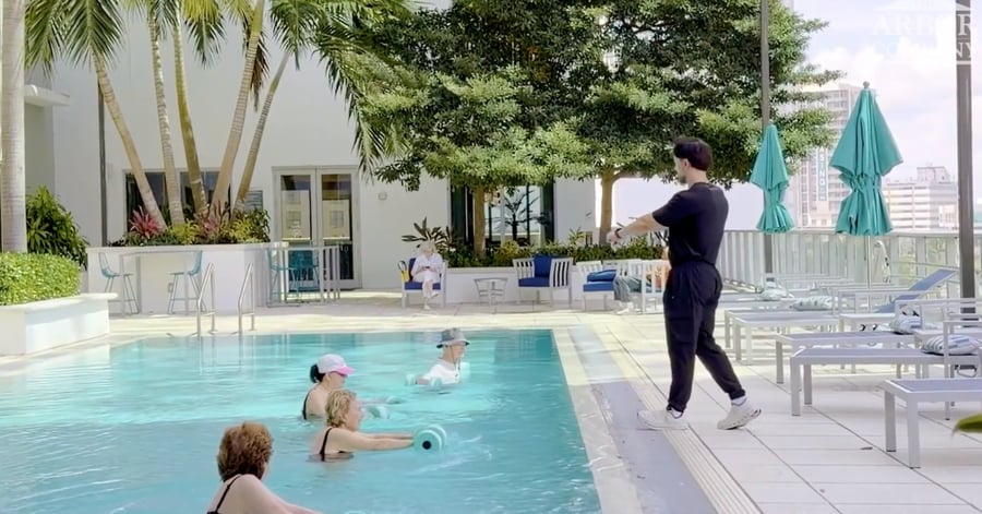 A rooftop pool deck where a fitness instructor in a black outfit stands poolside leading a water aerobics class. Four women stand in the pool holding foam dumbbells, wearing swim caps or sun hats. Behind them, lounge chairs, teal umbrellas, palm trees, and city high-rises are visible under a bright sky, with two seated onlookers in the background near the building.