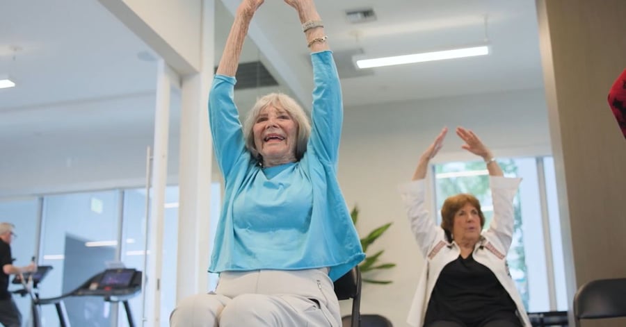 Two residents joyfully raise their arms during a seated fitness class, reflecting the active senior living and wellness programs at Mirabelle in Miami, FL.