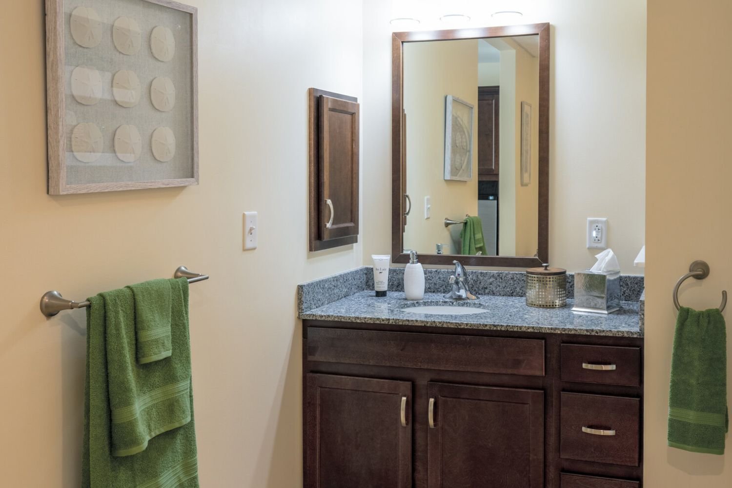A clean, well-appointed bathroom at this assisted living Shrewsbury NJ community, featuring a granite-top vanity, dark wood cabinetry, large mirror, and green accent towels.