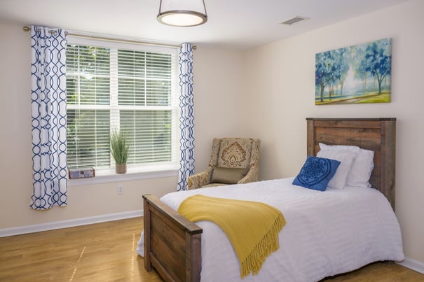 A cozy resident bedroom at this senior living Shrewsbury NJ community with a warm wood-frame bed, soft yellow throw, blue accent pillow, natural light from blinds, and a landscape painting on the wall.