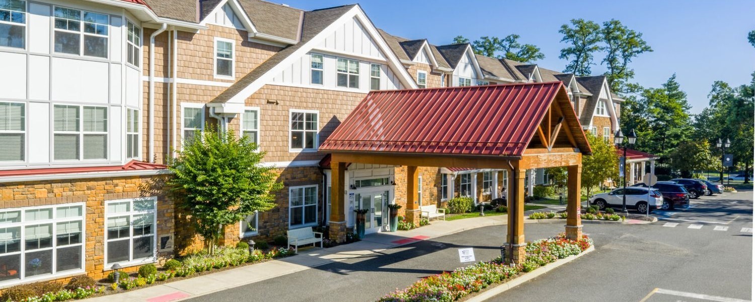 Exterior of a multi-story senior living community building featuring a covered porte-cochère entrance with a red metal roof and wooden timber frame, tan brick and shingle siding, white-trimmed windows, manicured landscaping with flowering plants and shrubs, and a paved parking area on a sunny day.