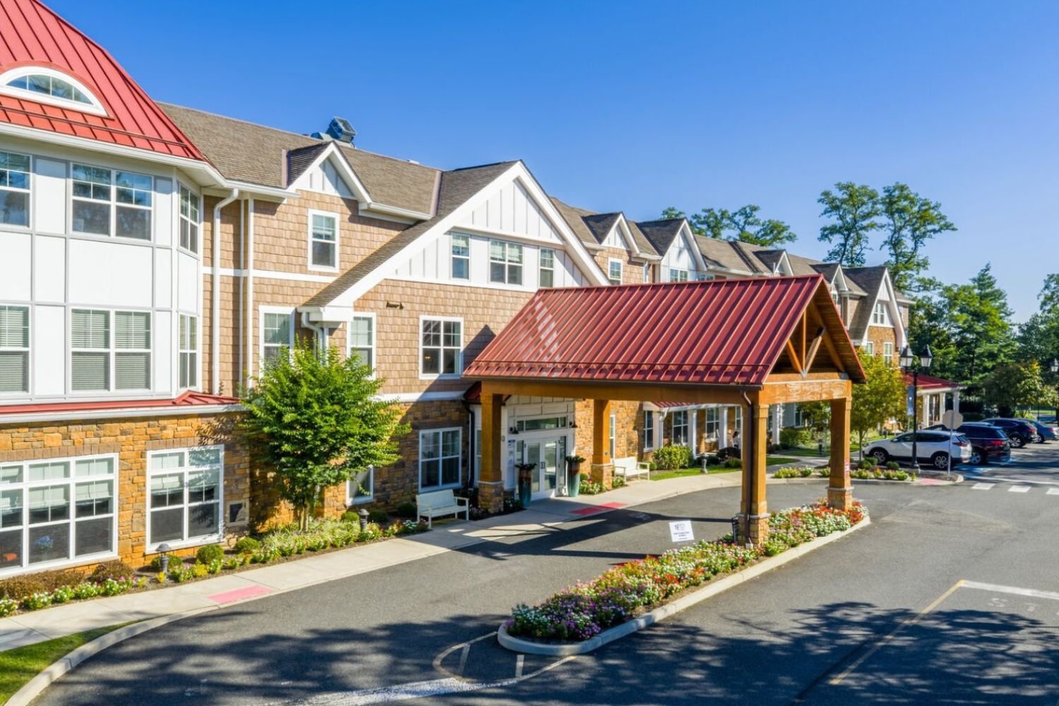 The welcoming front entrance of Arbor Terrace Monmouth, a senior living Shrewsbury NJ community, featuring a covered porte-cochère, red roof accents, lush landscaping, and a well-maintained arrival drive.