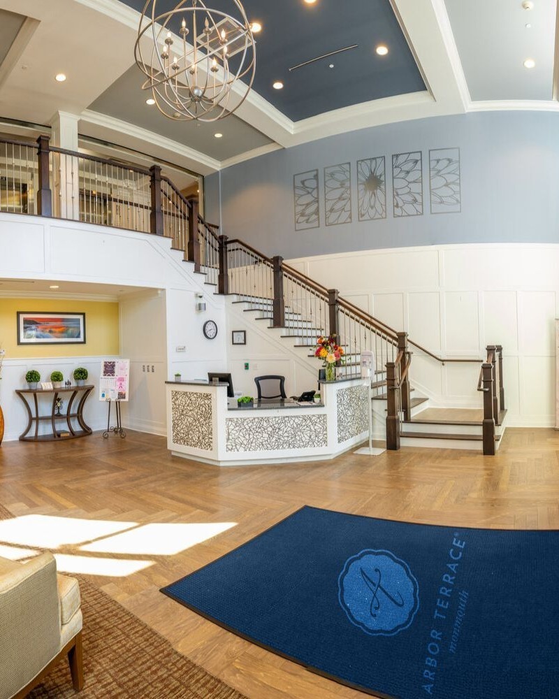 An elegant lobby with a sweeping staircase and reception area welcomes residents and visitors to this senior care Shrewsbury New Jersey community.