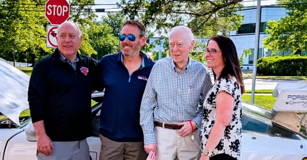 Four people smiling and posing together outdoors beside a white Porsche convertible. From left to right: an older man in a black New York Knicks pullover, a middle-aged man with a beard wearing blue mirrored sunglasses and a navy quarter-zip, an elderly man in a light blue checked button-down shirt and khakis, and a woman with dark hair and glasses in a white floral blouse. A stop sign and lush green trees are visible in the background