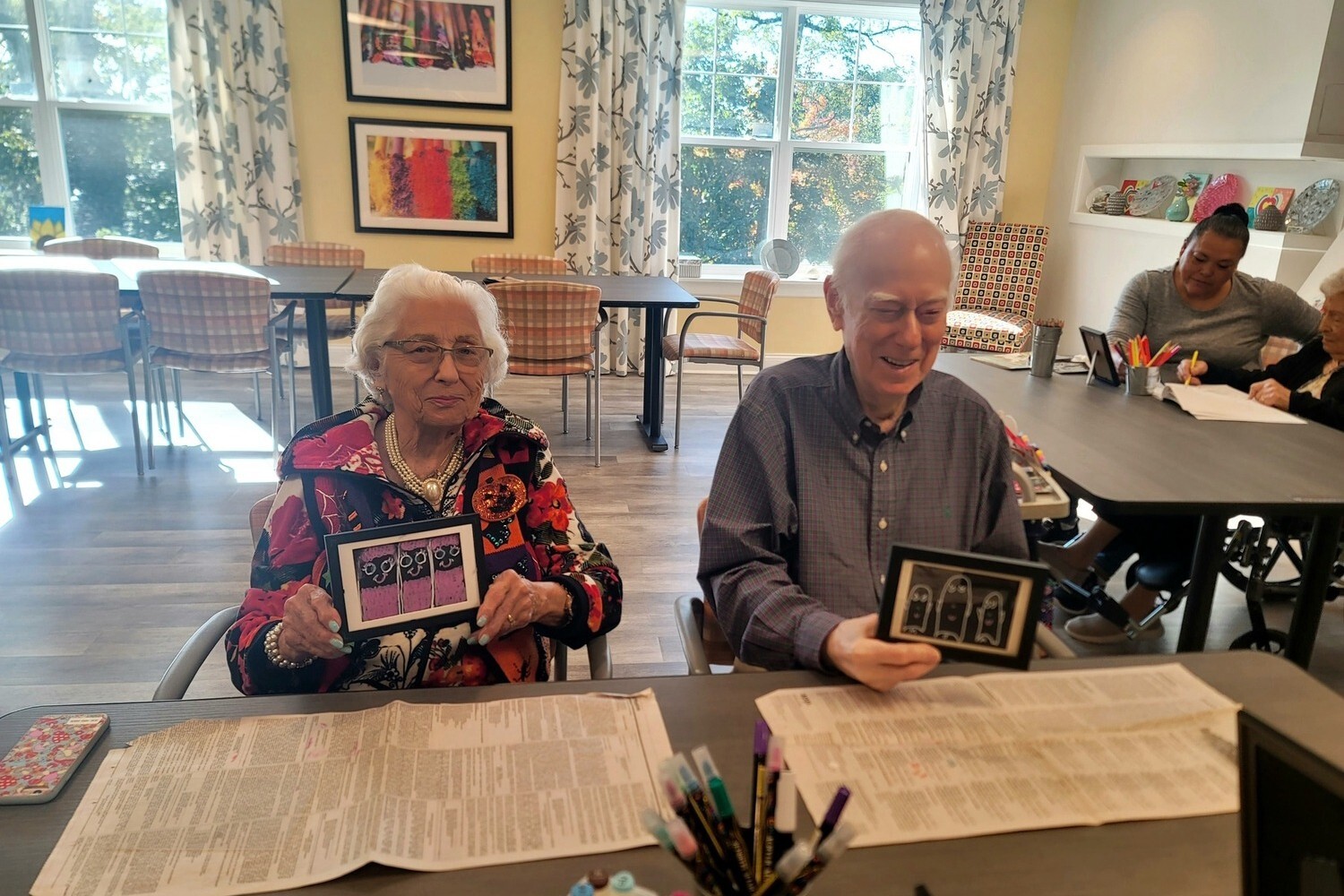 Two residents share a laugh while looking at photos together in a sunny lounge at Arbor Terrace Monmouth, a senior living community in Shrewsbury, NJ.