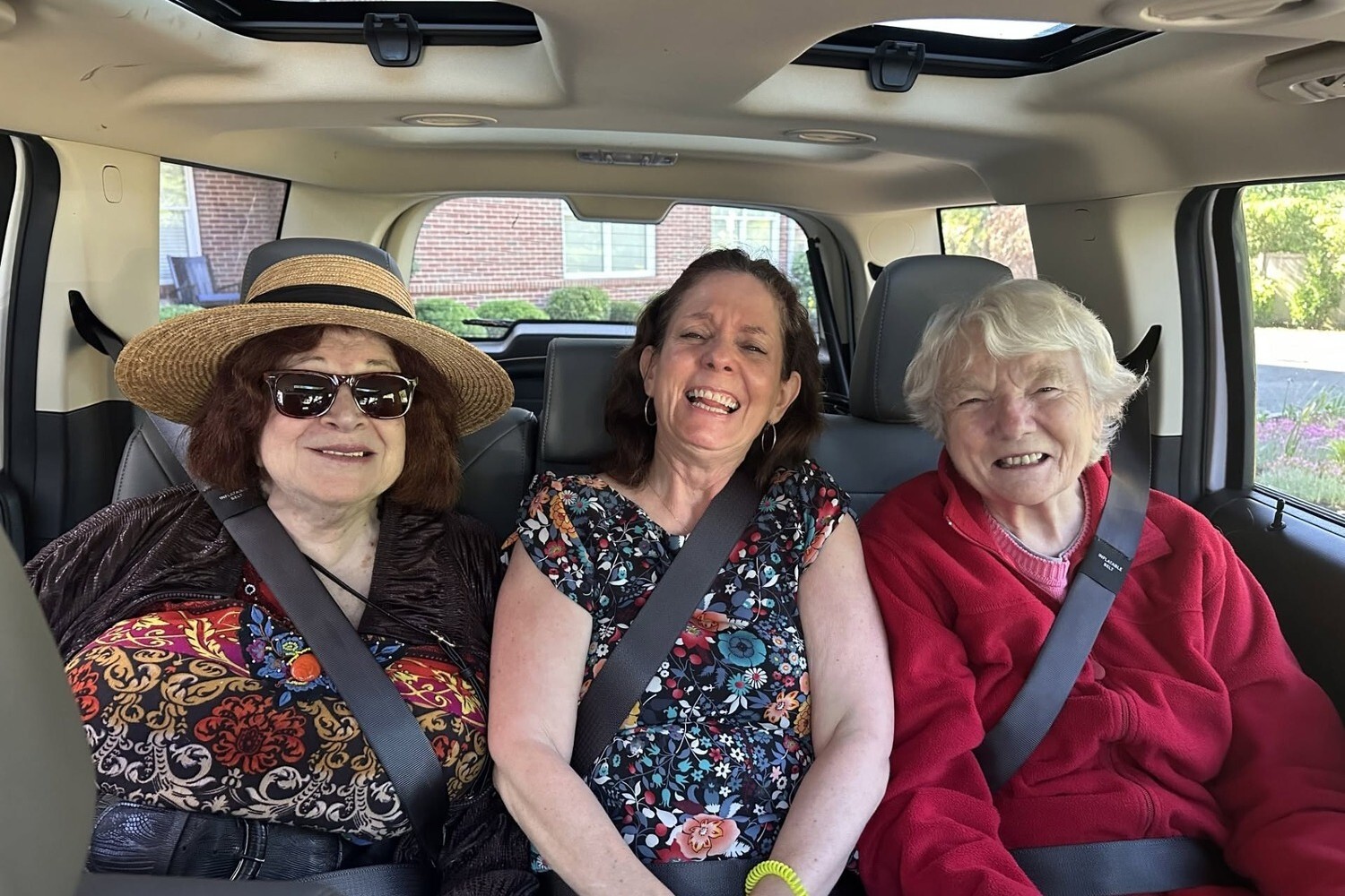 Three laughing women buckle up for a road trip, one in a straw hat and sunglasses, one in a floral dress, and one in a red fleece, a joyful ride that captures the spirit of active senior communities in Atlanta, GA and senior living near East Atlanta, GA.
