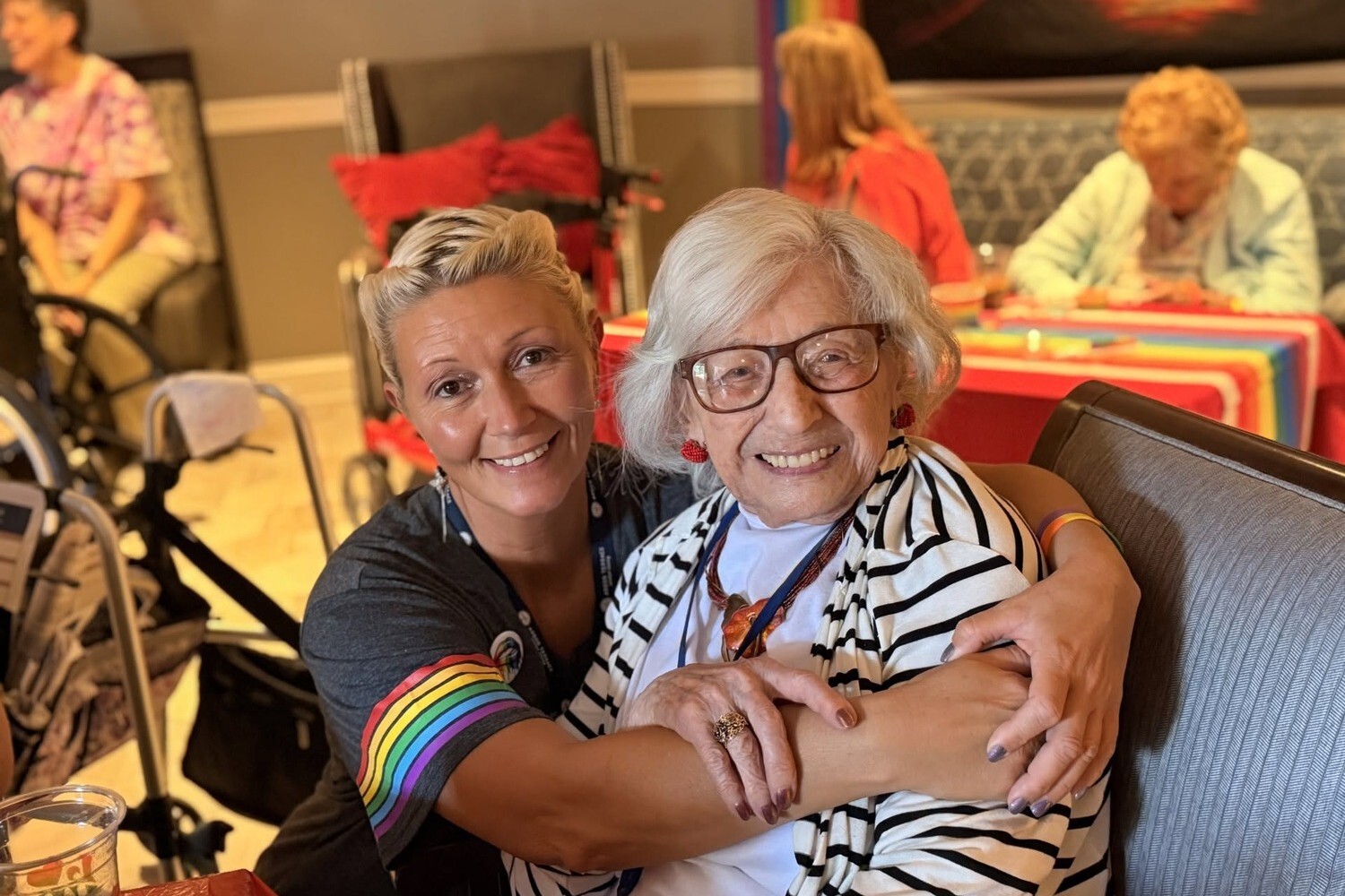 A smiling woman with blonde hair and a rainbow-striped sleeve wraps her arms around a senior woman with white hair, glasses, red earrings, and a striped cardigan, a heartfelt embrace that captures senior living in Atlanta, GA and senior living near East Atlanta, GA.