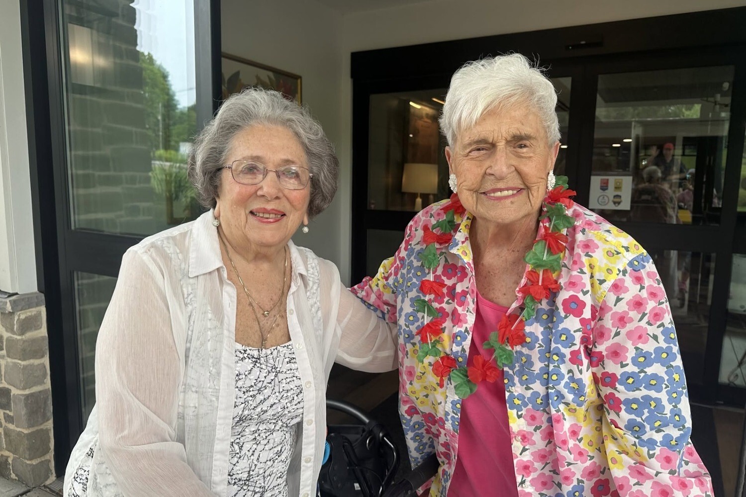 Two smiling senior women pose side by side, one with curly gray hair, glasses, and a sheer white blouse, the other with white hair, a bright floral shirt, and a festive red and green lei, a cheerful moment in senior living near East Atlanta, GA and retirement communities near Midtown Atlanta.