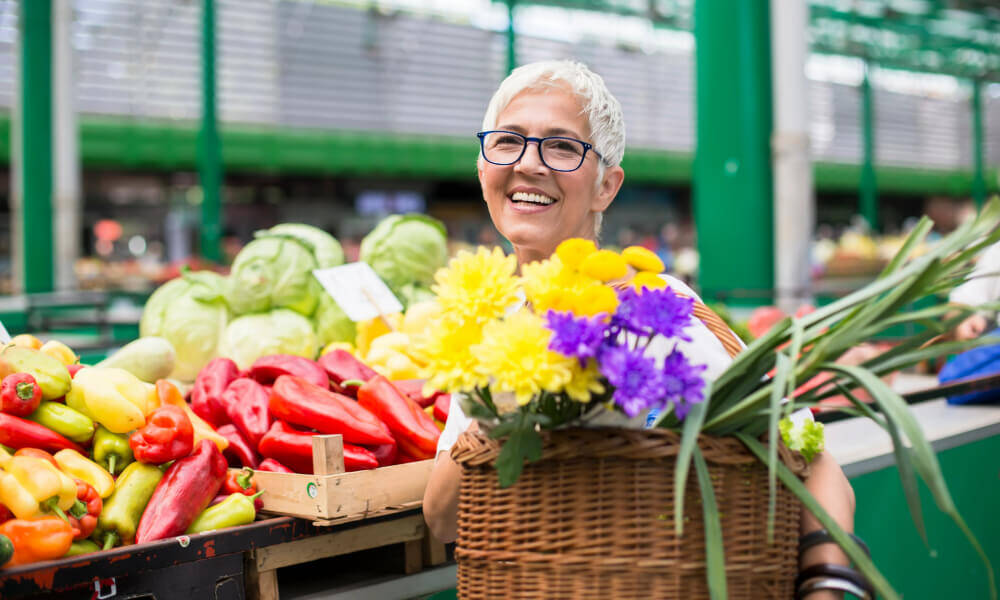 Senior woman shopping at a farmers market, smiling and holding a basket filled with fresh produce and flowers.