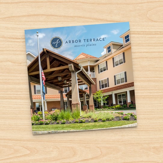 Square photo of Arbor Terrace Morris Plains building exterior with timber-framed entrance canopy, flagpole, and landscaped lawn, displayed on a printed card against a wood-grain background.
