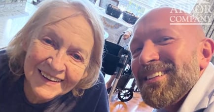 Selfie of a smiling older woman with gray hair and a bald, bearded man, taken indoors near a wheelchair and a sunlit window.