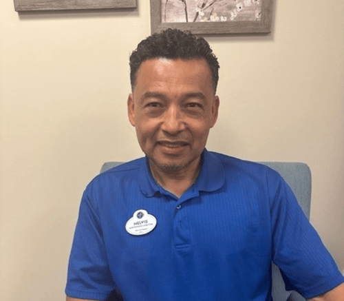 Headshot of Melvis, a team member at Arbor Terrace Morris Plains, wearing a blue shirt and nametag, smiling while seated.