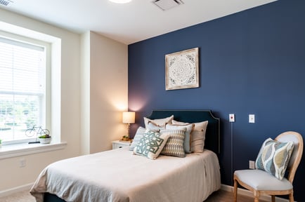 A warm, well-appointed bedroom at this assisted living community near Mount Laurel NJ, featuring a bold navy accent wall, a neatly dressed bed with layered pillows, a bedside lamp, and a bright window with views of greenery.
