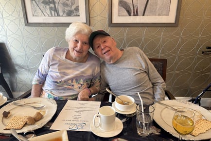 Two smiling seniors lean in close at a dining table set with silver-rimmed plates, soup, and coffee, sharing a warm moment over a meal that reflects the connection at the heart of wellness-focused senior living South Jersey and an active senior lifestyle Mount Laurel NJ.