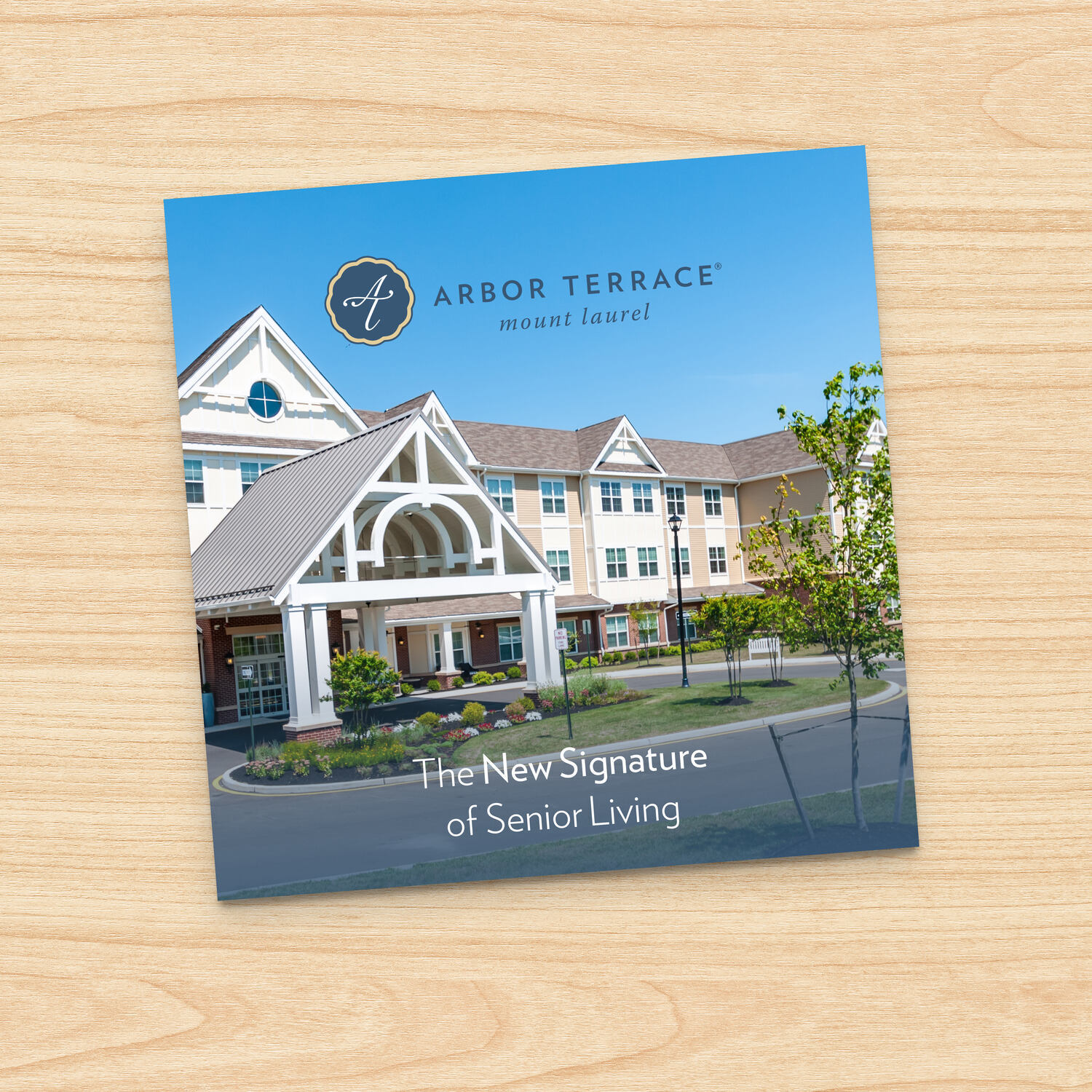 Guide cover for Arbor Terrace Mount Laurel resting on a light wood surface, showing the covered front entrance and fresh landscaping under a blue sky. A look at senior living in Mount Laurel, NJ, and senior care across Burlington County.