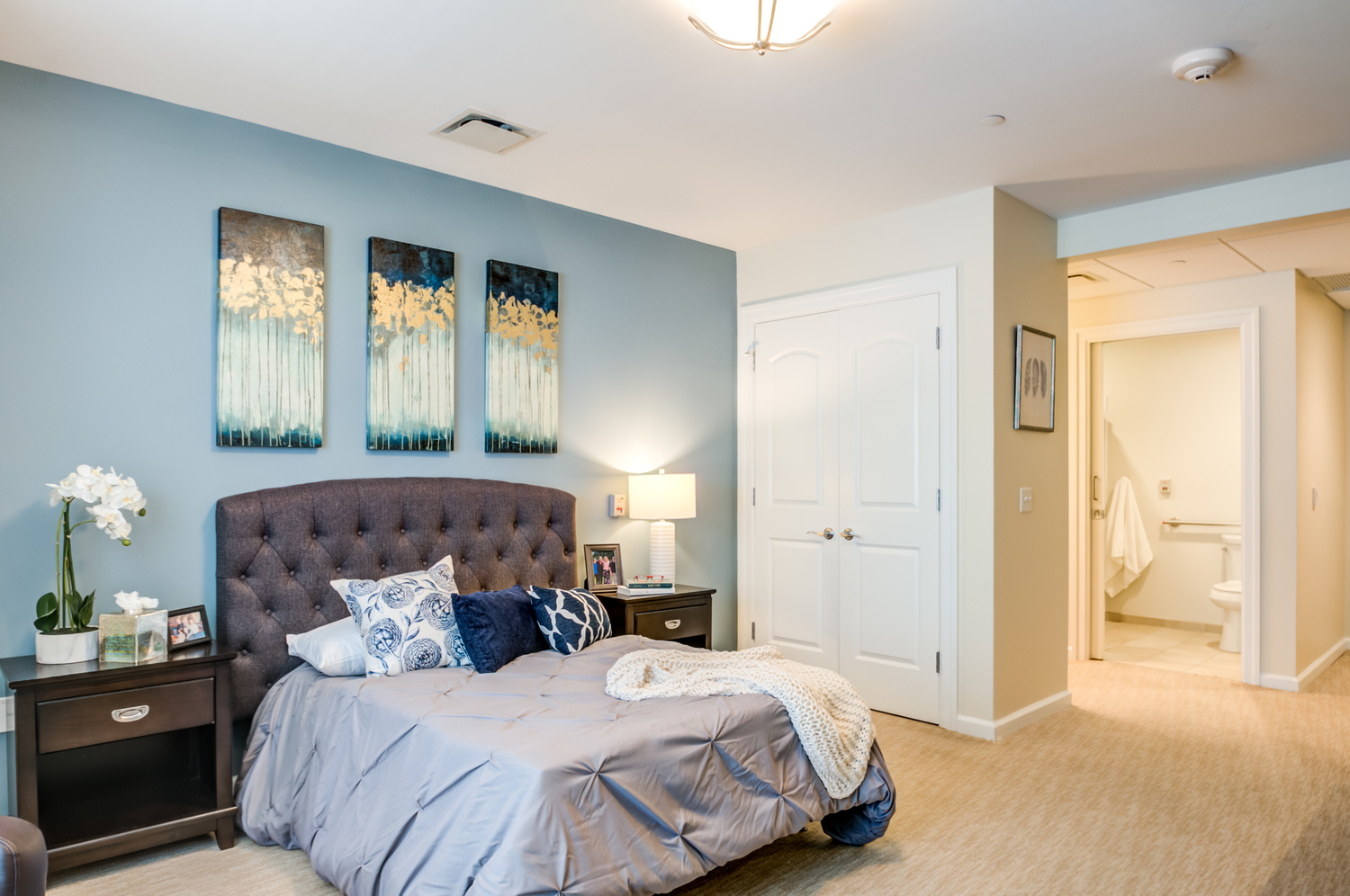 A spacious apartment bedroom at our senior living Mountainside NJ community features a tufted headboard, layered artwork, and a private ensuite bathroom, reflecting the comfortable, active senior lifestyle North Jersey residents enjoy.