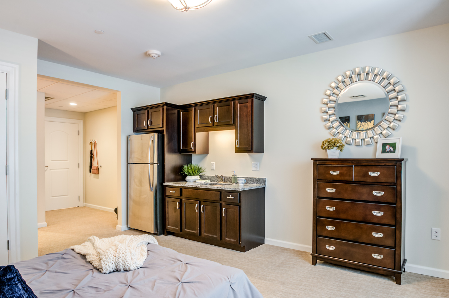 A convenient kitchenette in an apartment at our senior living Mountainside NJ community features rich wood cabinetry, a full-size refrigerator, and a stylish mirrored accent, reflecting the wellness-focused senior living Mountainside lifestyle.