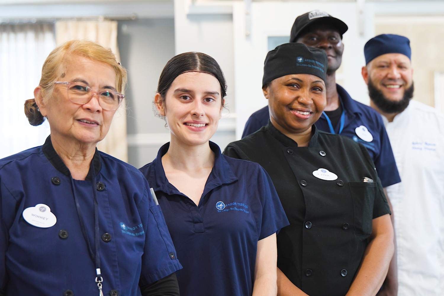 Five smiling culinary and support team members pose together warmly in their kitchen uniforms, reflecting the dedication and camaraderie of the senior living team within this senior living community.
