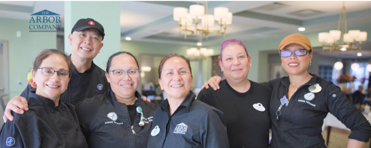 Six team members from The Arbor Company share a joyful embrace at a senior living celebration, smiling in matching branded polos within a chandelier-lit banquet room, reflecting the warmth of this active senior living team.
