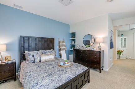 A spacious, well-appointed bedroom at this assisted living community in Norwood NJ, featuring a dark wood headboard and dresser, soft blue accent wall, a round mirror, built-in shelving, and an open doorway leading to an adjoining room.