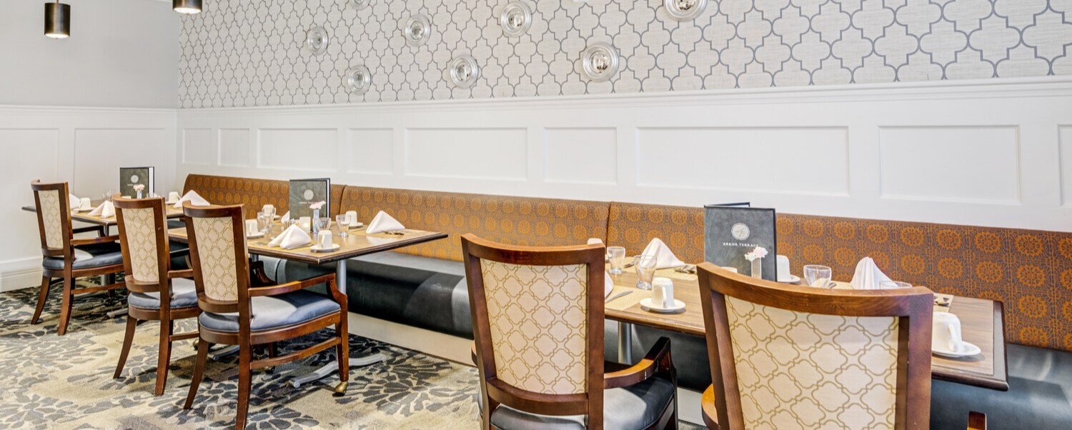 Elegantly set dining tables with upholstered chairs, booth seating, and patterned wallpaper in the dining room at Arbor Terrace Norwood, a welcoming assisted living community in Norwood, NJ.