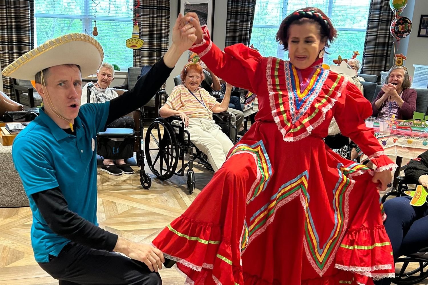 residents at Arbor Terrace Norwood enjoy a lively Cinco de Mayo celebration, with a performer in traditional dress holding hands with a staff member in a sombrero. In the background, residents in wheelchairs/chairs enjoy the festive atmosphere with colorful decorations