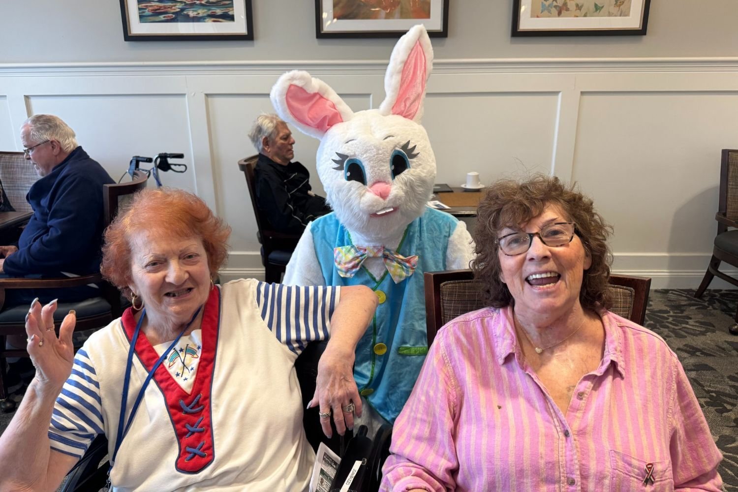Two smiling residents sit and pose with the Easter Bunny mascot at Arbor Terrace Norwood. One resident wears a red, white, and blue shirt, and the other wears a pink striped shirt. In the background, other residents sit at tables