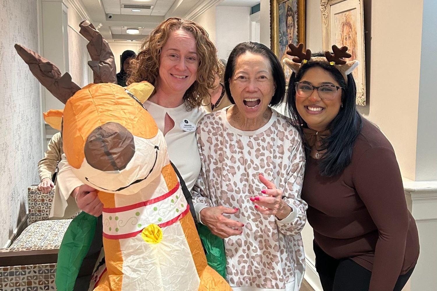 Three women smile and pose together at Arbor Terrace Norwood during a festive holiday event. One of the women is holding a large inflatable reindeer, another is wearing  reindeer antlers, and the other in a leopard-print sweater.