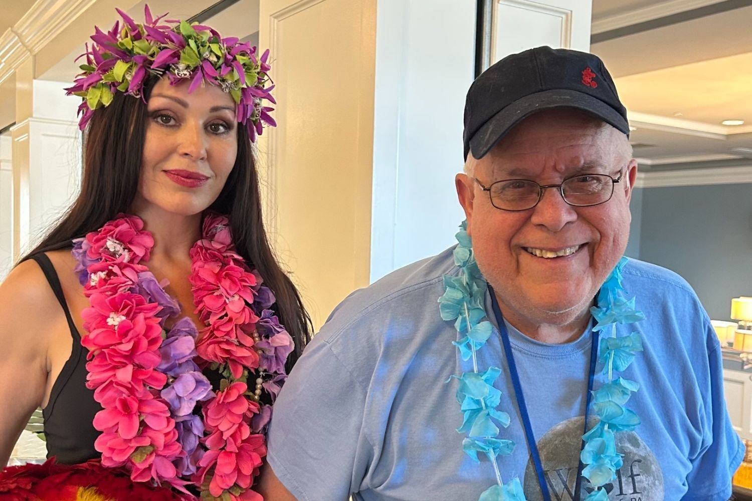 A man and a woman are smiling and standing together at Arbor Terrace Norwood during a hula dance social. The woman is dressed in vibrant flower leis, while the man wears a casual shirt, glasses, a blue lei, and a hat