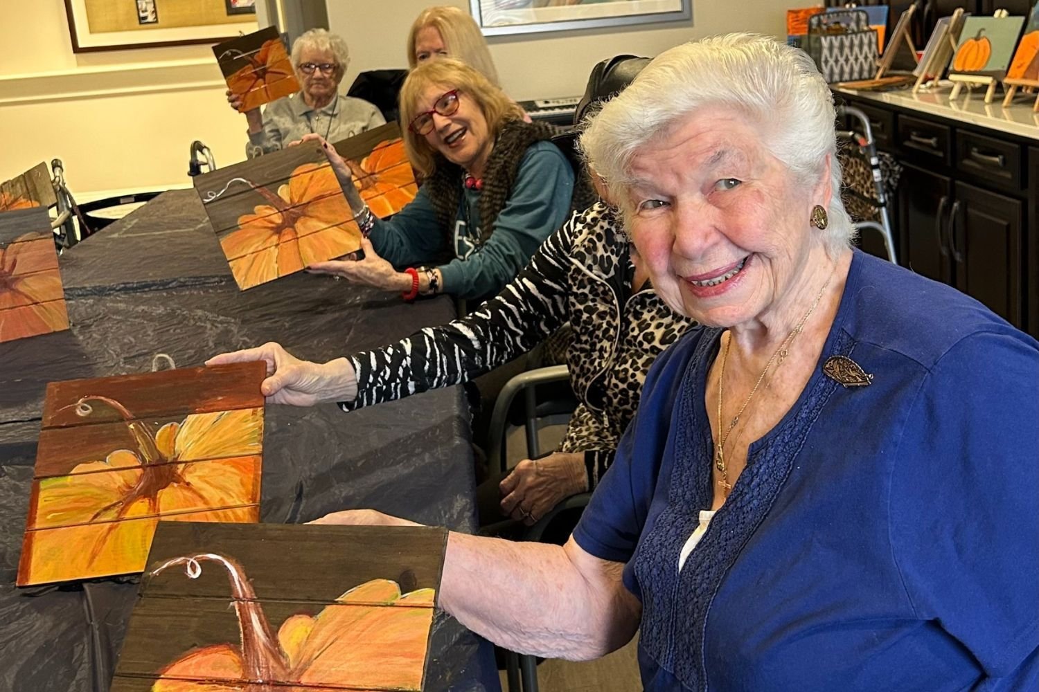A group of residents at Arbor Terrace Norwood gather around a table and each show off a painting of a pumpkin. The residents smile