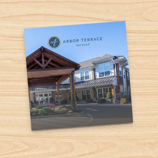 Square photo of Arbor Terrace Norwood building exterior with timber-framed entrance canopy and multi-story facade, displayed on a printed card against a wood-grain background.