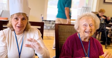 Two elderly women sit together at a table in what appears to be a senior living community dining room, smiling and laughing. The woman on the left wears a white paper chef's hat, a blue lanyard, and a disposable glove, gesturing with her hand while speaking. In front of each woman is a small white ramekin bowl filled with a colorful food dish. The woman on the right, wearing a maroon sweater and a blue lanyard, holds a cracker and smiles warmly at the camera. In the background, a staff member in a teal shirt stands near a window, and another resident is visible at a table in the rear.
