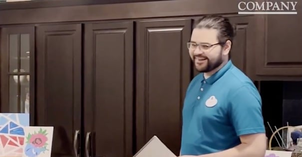 A smiling man with glasses and a beard wearing a blue polo shirt with a name badge stands in front of dark wood cabinetry, with colorful artwork visible in the foreground.