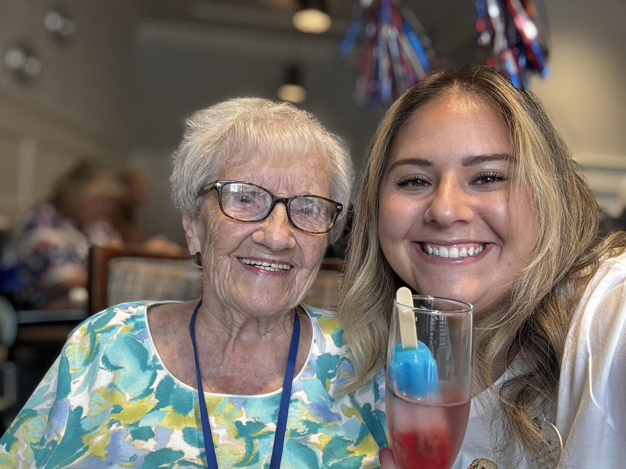 A resident and a staff member smile together at a festive celebration, holding colorful drinks at Arbor Terrace Norwood, a warm and engaging senior living community in Norwood, NJ.