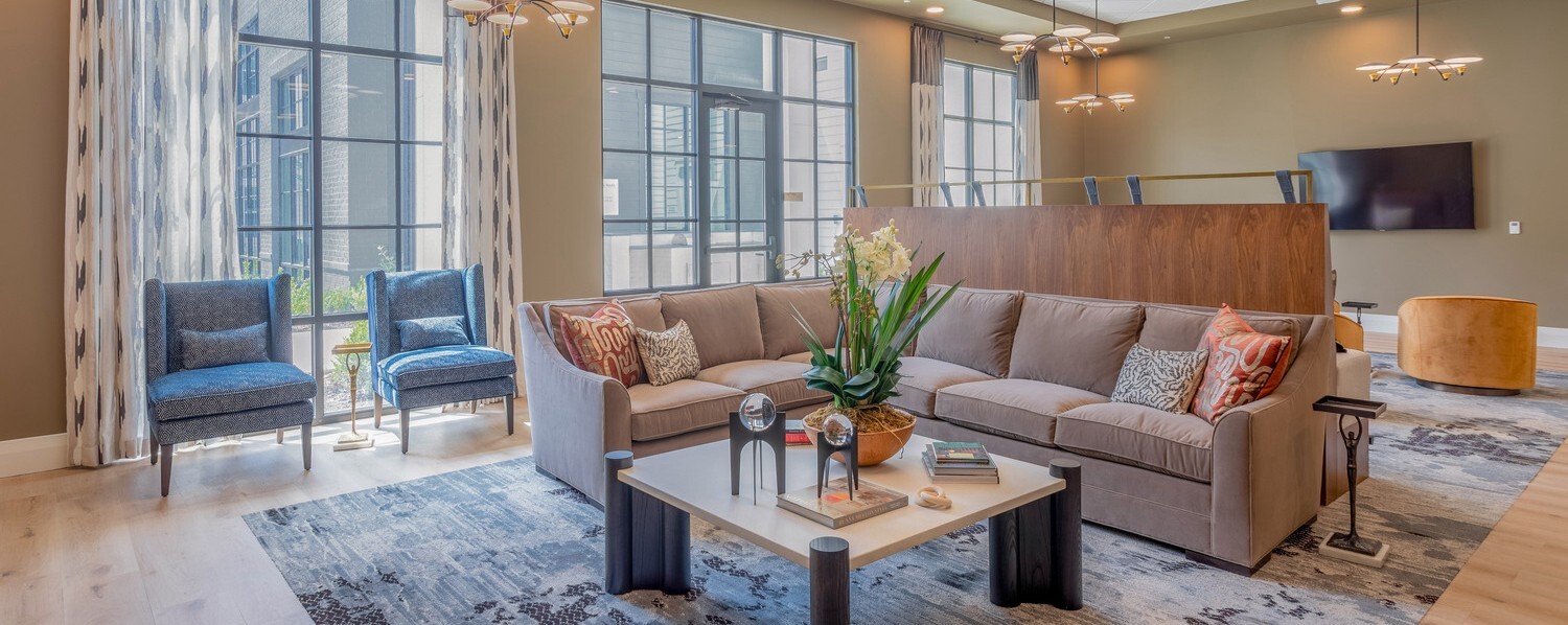 A bright, welcoming common area at Opus East Memphis senior living in East Memphis, featuring a large sectional sofa with colorful throw pillows, blue accent chairs, and floor-to-ceiling windows that flood the space with natural light.