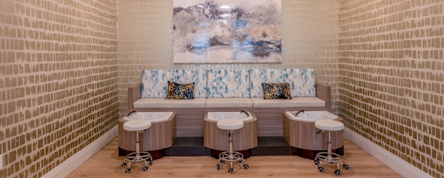A spa pedicure suite with three wooden pedicure stations, white cushioned technician stools on wheels, a built-in bench with blue and white patterned upholstery and decorative pillows, textured tan wallpaper, light hardwood flooring, and an abstract landscape painting mounted on the wall above the bench