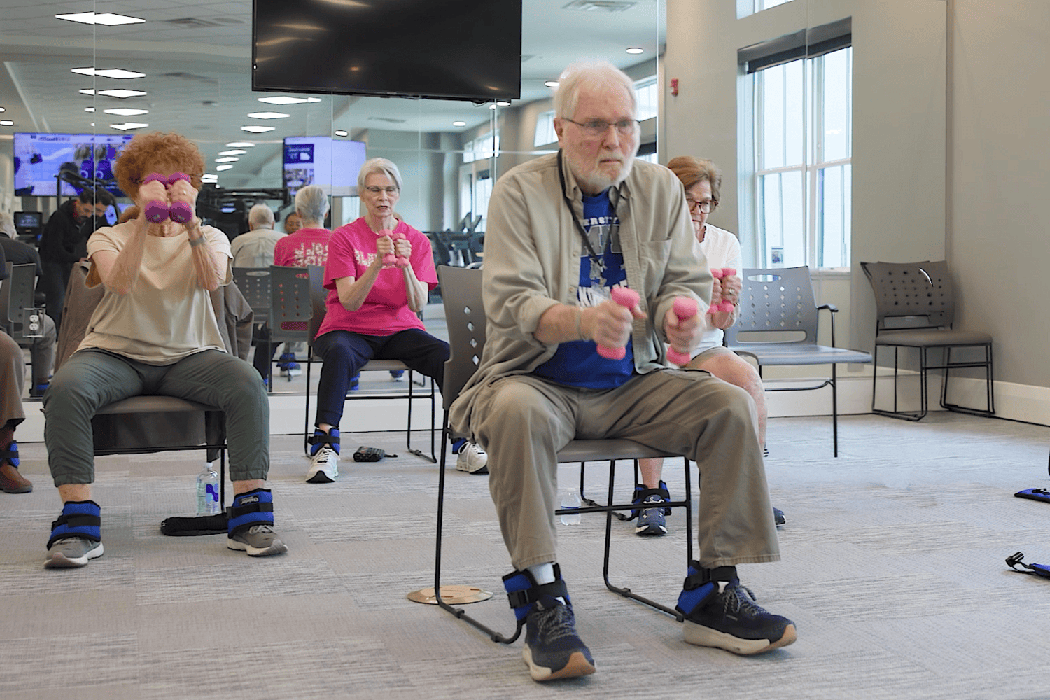 Seated side by side in a bright exercise room, a group of seniors lifts pink hand weights, including a man in glasses and a khaki shirt and a woman in a pink tee, an energizing class in active senior communities in Atlanta, GA and top-rated senior living in Atlanta, GA.