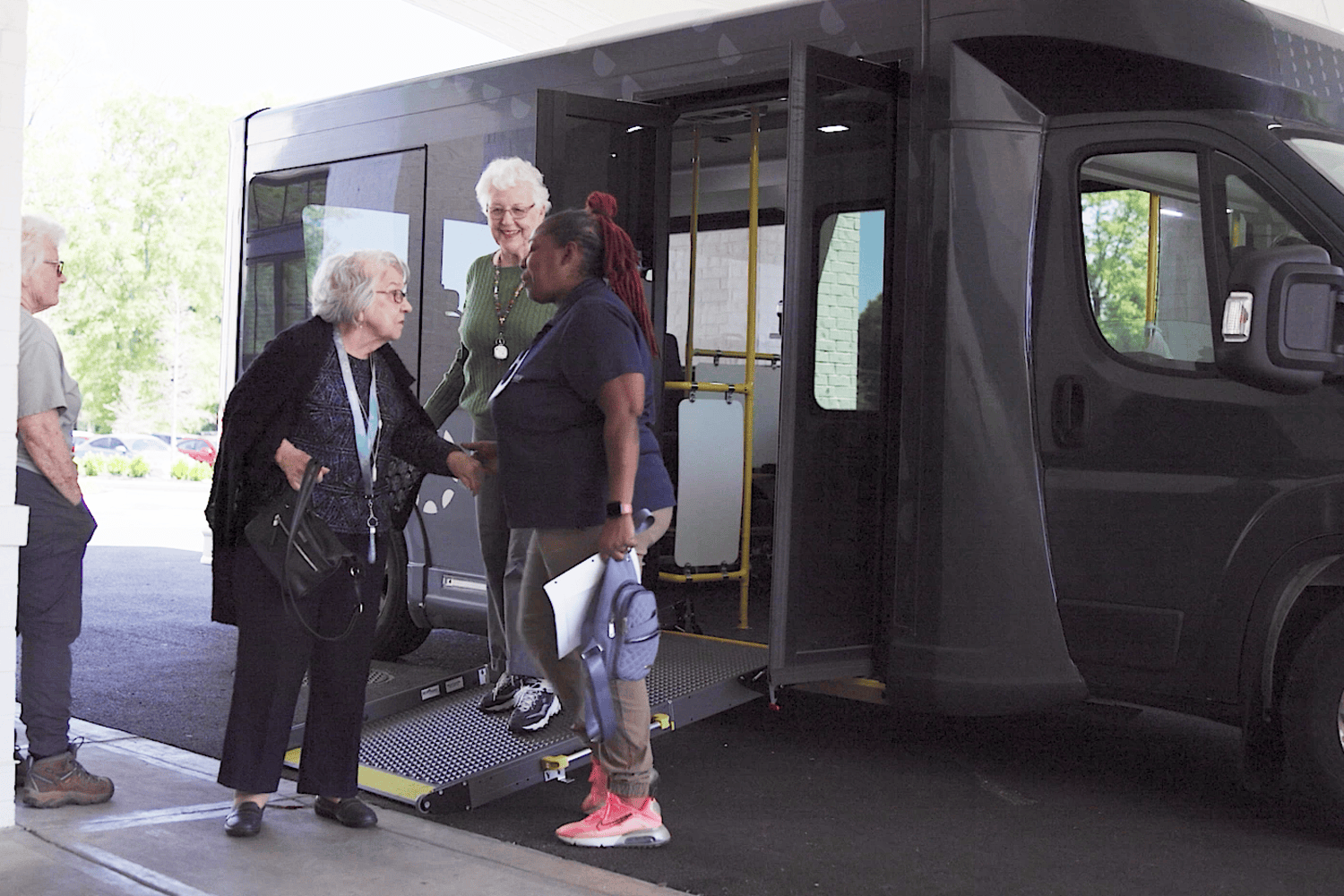 Two senior women, one in a green sweater and one in a dark cardigan, chat with a woman in a navy polo at the door of a shuttle bus, a friendly moment that reflects active senior living Atlanta, GA and senior living near Buckhead, GA.