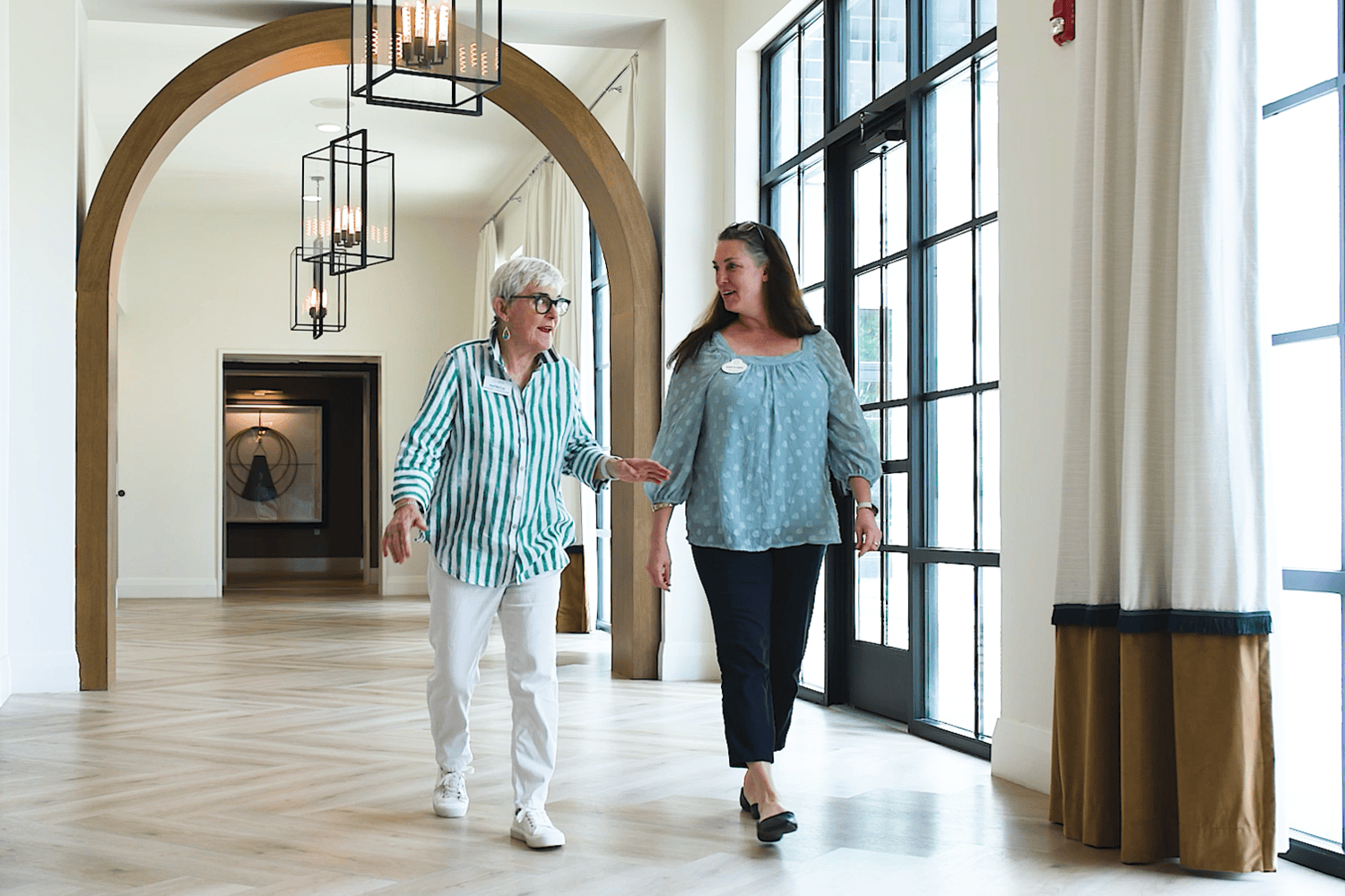 Two women chat while strolling through a bright hallway with a wooden archway, lantern pendant lights, and tall black-framed windows, one in a green striped shirt, the other in a soft blue blouse, a welcoming look at senior living in Atlanta, GA and senior housing near Buckhead, GA.