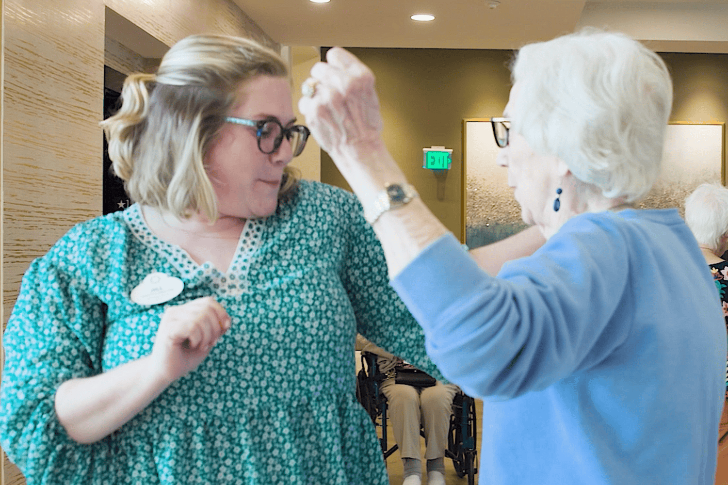 A woman with blonde hair, glasses, and a green floral dress dances alongside a senior woman with white hair and a blue sweater who raises her arm with flair, a playful moment of senior living in Atlanta, GA and senior living near Midtown Atlanta, GA with social activities.
