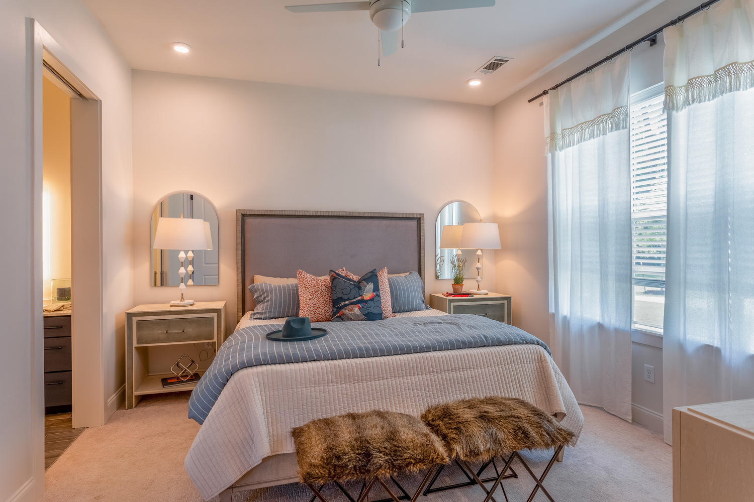 A serene, elegantly styled bedroom at this senior living community in Memphis TN, featuring a tall upholstered headboard, coordinating nightstands with arched mirrors, soft blue bedding, faux fur accent stools, a ceiling fan, and sheer curtains letting in warm natural light.