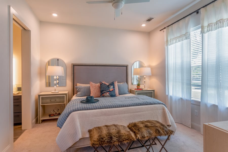 A serene, elegantly styled bedroom at this senior living community in Memphis TN, featuring a tall upholstered headboard, coordinating nightstands with arched mirrors, soft blue bedding, faux fur accent stools, a ceiling fan, and sheer curtains letting in warm natural light.