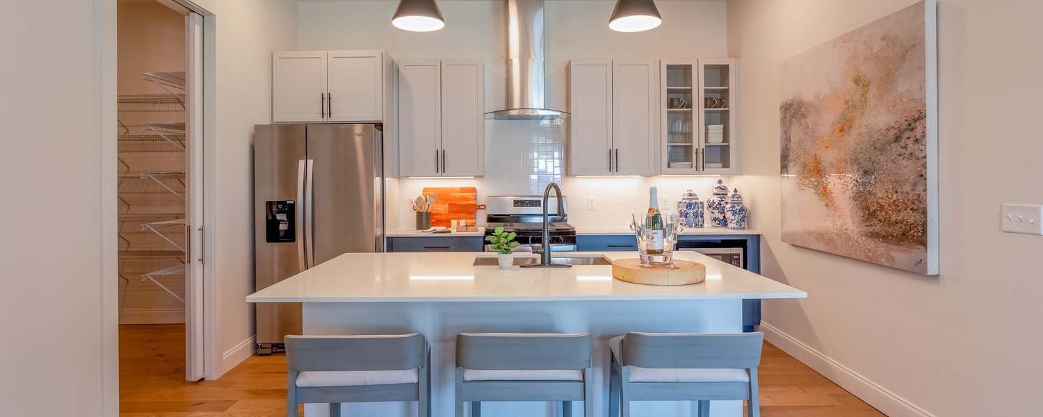 Modern kitchen with white cabinetry, a large island with white countertop and three gray bar stools, stainless steel appliances, a stainless range hood, pendant lighting, and decorative blue and white ceramic vases on the counter, with light wood flooring and a walk-in pantry visible to the left.